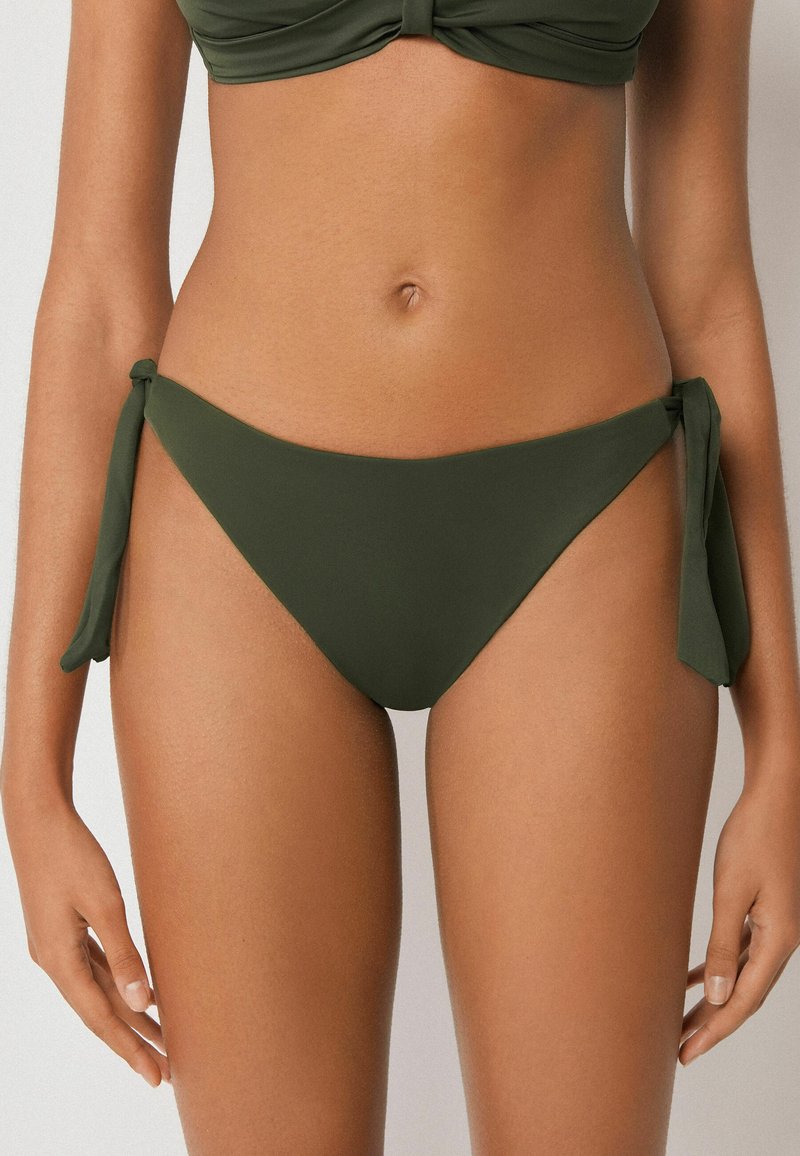 Frau trägt olivegrünen Bikinihöschen mit seitlichen Schnürungen, zeigt den Bauchbereich und die Hände vor neutralem Hintergrund.