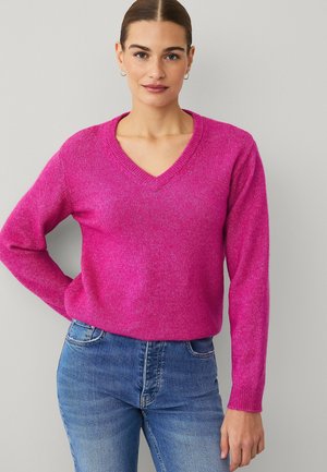 Vrouw met naar achteren gestyled haar, zilveren creolen, een felroze V-hals trui en een blauwe spijkerbroek, staand tegen een lichtgrijze muur.