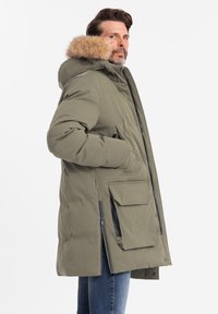 Olivgrüne Steppjacke mit Kapuze aus Kunstfell, mehreren Taschen und einem Reißverschluss vorne. Sie verfügt über ein gestepptes Design und verstellbare Ärmelbündchen.