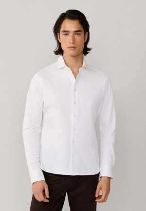 Muž so stredne dlhými tmavými vlasmi, oblečený v bielej košeli na gombíky a tmavých nohaviciach, na jednoduchom svetlom pozadí.