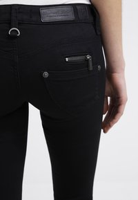 Jeans de mezclilla negra con un bolsillo trasero con cremallera, diseño de yugo curvado y una etiqueta de cuero en la cinturilla.