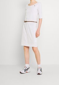Robe grise clair midi avec un col rond, des manches 3/4, une ceinture tressée marron et un petit détail de logo. Associée à des baskets blanches sportives.