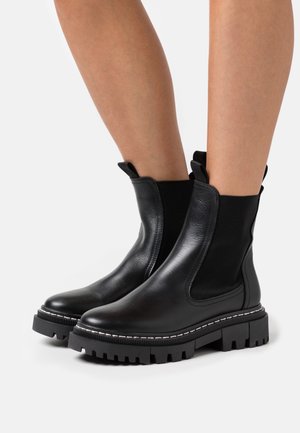 Bottes en cuir noir montant à la cheville avec semelles épaisses à crampons, portées sur des jambes nues, avec des panneaux élastiques sur les côtés et des languettes pour les enfiler.
