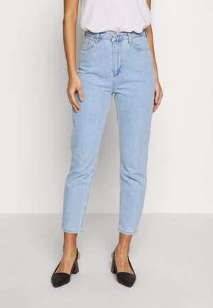 Femme portant un jean taille haute à jambes droites bleu clair, un haut blanc et des chaussures à talons carrés noires à bride arrière, debout devant un fond uni.