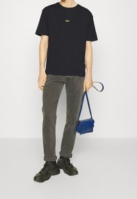 Svart bomullströja med en gul logotyp, grå byxor, svarta sneakers och en liten blå handväska med rem.