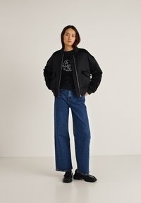 Čierna bomber bunda s lesklým povrchom, nosená cez čierne tričko s potlačou. Kombinovaná so širokými modrými džínsami a čiernymi topánkami.