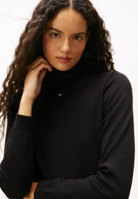 Donna con capelli ricci e scuri che indossa un maglione nero a collo alto, appoggiando una mano vicino al mento, guardando dritto davanti a sé.