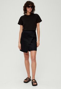 Zwarte katoenen t-shirt gecombineerd met een zwarte wikkelrok met knopen, textuurstof en platte zwarte sandalen. Model draagt oversized zonnebril.