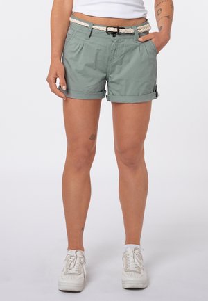 Person trägt gerollte grüne Shorts mit einem weißen geflochtenen Gürtel, weißen Turnschuhen und einem Teil eines weißen Tops und steht mit den Händen nahe den Taschen.