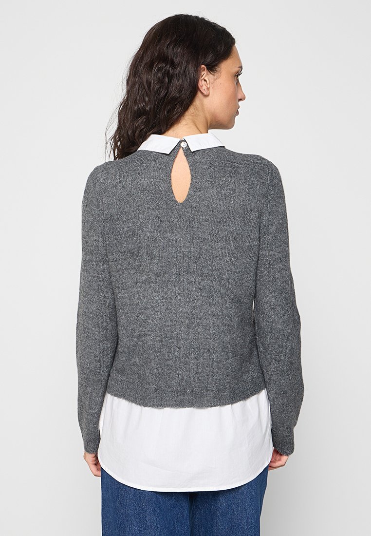 Grauer Strickpullover mit langen Ärmeln, Schlüsselriegel-Detail hinten und einem weißen Kragenhemd darunter, kombiniert mit blauen Jeans.