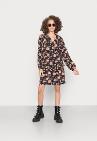 Vestido floral com mangas compridas, base preta, flores cor-de-rosa e laranja, decote em V. O calçado apresenta botas de tornozelo pretas com fivela.
