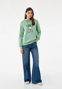 Felpa verde menta con stampa grafica, tasca a marsupio anteriore e cappuccio con cordino, abbinata a jeans larghi blu e scarpe di colore chiaro.