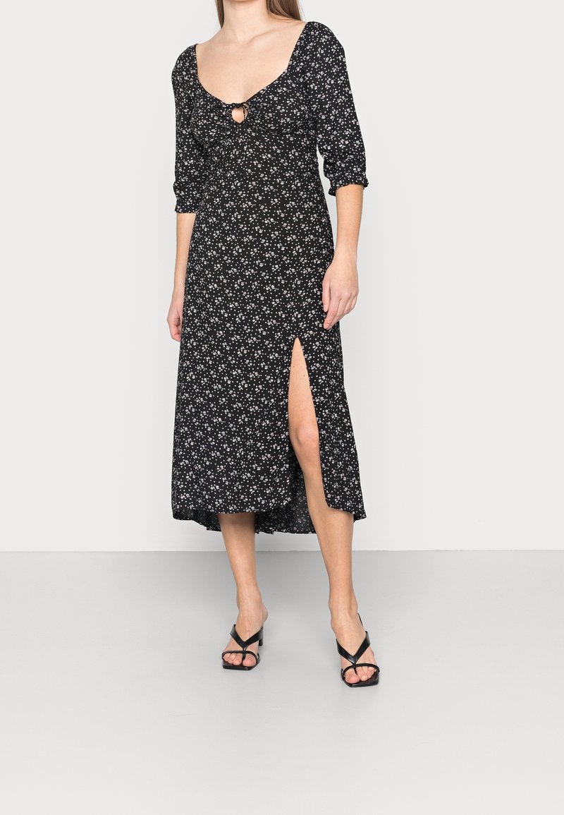 Femme portant une robe midi noire à petit imprimé floral blanc, manches trois-quarts, un keyhole à l'avant et une fente haute à la cuisse, associée à des talons noirs.