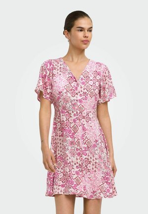 Kvinde iført en kort kjole med pink og hvide blomstermønstre, med flyveærmer og V-udskæring, stående mod en ensfarvet baggrund.