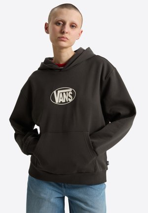 Person med kort hår iført en mørk Vans hættetrøje og blå jeans, stående med hænderne i lommerne mod en ensfarvet baggrund.