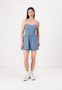 Denim obleka z ozkimi naramnicami, oprijetim korpusom in širokim krilom. Svetlo modra barva, mehak površinski sloj in vidne šive.