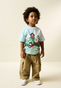 Tricou din bumbac albastru deschis cu broderie colorată cu tematică agricolă, asortat cu pantaloni cargo bej și adidași albi. Mâneci scurte și croială lejeră.