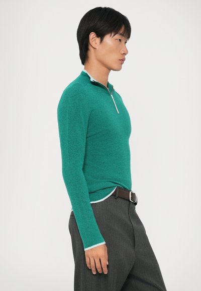 Pullover verde, texturizado, com fecho de zip e com detalhes brancos contrastantes na gola e nos punhos, combinado com calças cinzentas riscas e um cinto castanho.