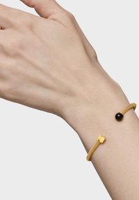 Złota bransoletka z siateczki z czarnym koralikiem i złotym akcentem w kształcie motyla. Noszona na dłoni z widoczną fakturą skóry.