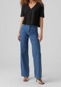 Czarny krótki top na guziki z krótkimi rękawami, zestawiony z jasnoniebieskimi jeansami o wysokim stanie i szerokich nogawkach. Modelka nosi czarne sandały na obcasie.