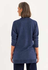 Frau mit schulterlangem Haar, die einen langärmligen, marineblauen Pullover und dunkle Hosen trägt, steht mit dem Rücken zu einem einfarbigen Hintergrund.