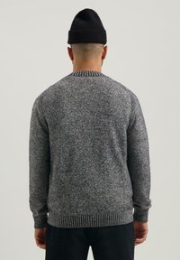 Hombre de espaldas vistiendo un suéter de punto gris oscuro, pantalones negros y un gorro negro, contra un fondo claro y liso.