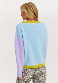Kolorowy, lekki sweter z niebieską bazą, fioletowymi rękawami i jasnobrązowym kołnierzykiem. Miękka struktura, luźny krój, warstwowy design.