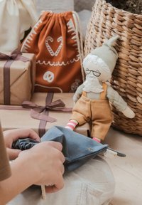 Muñeco gnomo de tela suave con un sombrero gris y barba blanca, vestido con pantalones naranjas. Cerca hay cajas envueltas para regalo y una bolsa de cordón con patrones.