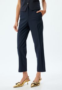 Pantaloni navy con una vestibilità su misura, caratterizzati da due tasche anteriori e una texture liscia. Abbinati a décolleté oro lucido con un dettaglio a fibbia.