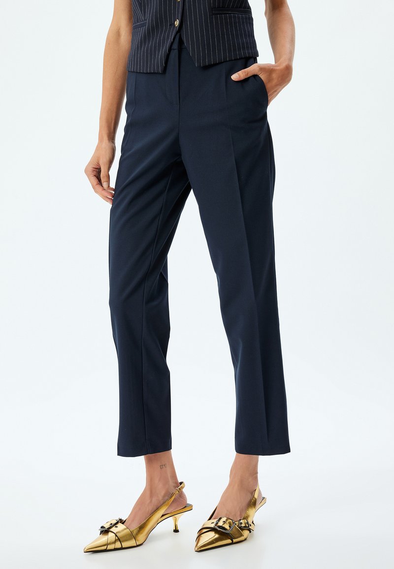 Pantaloni navy con una vestibilità su misura, caratterizzati da due tasche anteriori e una texture liscia. Abbinati a décolleté oro lucido con un dettaglio a fibbia.