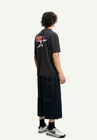 T-shirt in cotone nero con grafica rossa "05" e stella bianca sulla schiena, abbinata a pantaloni corti in denim blu scuro fino al ginocchio e scarpe da ginnastica argentate.