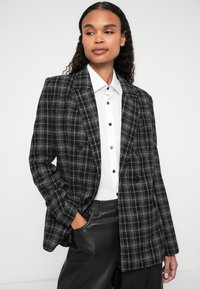 Dvoredni sako s crno-bijelim kariranim uzorkom izrađeno od teksturirane tkanine, nosi se preko bijele košulje s plavim gumbima i crnim kožnim hlačama.