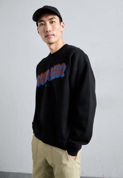 Schwarzer Sweatshirt mit der Aufschrift "SQUARED2" in blauer und roter Schrift. Verfügt über einen runden Ausschnitt und lange Ärmel. Kombiniert mit khakifarbenen Hosen.