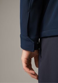Primo piano della mano di una persona rilassata lungo il lato del corpo, che indossa una giacca blu navy con polsino abbottonato e pantaloni blu navy abbinati.