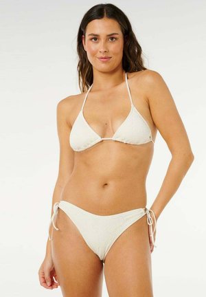 Donna che indossa un bikini bianco in crochet con top a collo allacciato e slip con fiocchi laterali, in piedi su uno sfondo bianco uniforme.