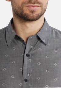 Camicia button-up grigia con piccoli motivi a logo grigio chiaro. Caratterizzata da un colletto classico, una texture sottile e due bottoni blu scuro sul davanti.