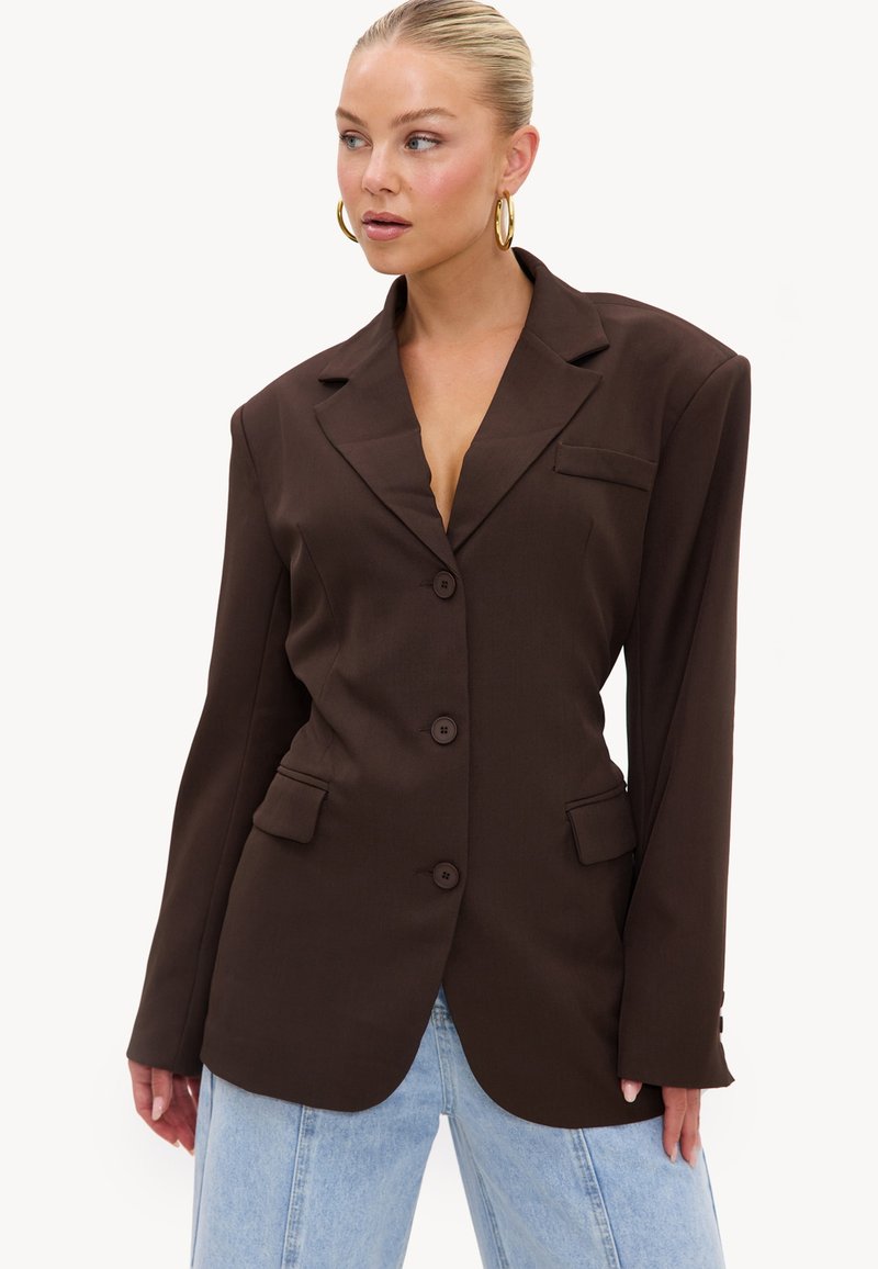 Femme aux cheveux blonds avec des boucles d'oreilles créoles dorées portant un blazer marron foncé oversize et un jean bleu clair sur fond blanc.