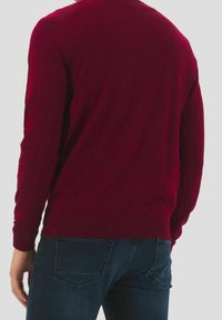 Maglione a maglia bordeaux con maniche lunghe, polsini e orlo a coste; texture morbida. Indossato con jeans di denim blu scuro.