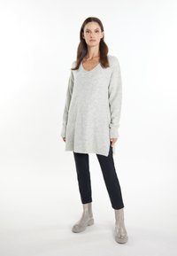 Pulover tricotat de culoare gri deschis, cu guler în V, mâneci lungi și șlițuri laterale, purtat cu pantaloni negri slim-fit și botine gri până la gleznă.