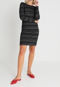 Kobieta w dopasowanej, czarnej sukience z długim rękawem w wzór zygzakowy, z czerwonymi płaskimi butami w czubek, stojąca na jednolitym tle.