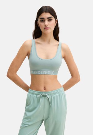 Frau mit langen dunklen Haaren, die ein hellblaues Sport-BH von Marc O’Polo und passende Jogginghosen trägt, steht mit den Händen im Rücken.