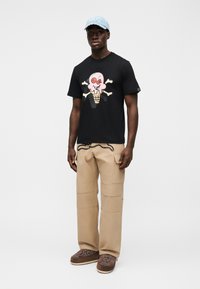 Μαύρο T-shirt με πολύχρωμο γραφικό ενός χωνάκι παγωτού και οστών, συνδυασμένο με μπεζ παντελόνι cargo και ανοιχτό μπλε καπέλο με κέντημα.