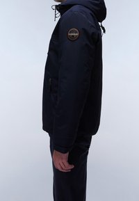 Granatowa kurtka z kapturem i regulowanym sznurkiem, z naszywką na górnej części rękawa, kieszenią na zamek po boku oraz gładką teksturą.