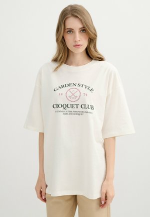 Joven mujer con cabello rubio largo que lleva una camiseta blanca de gran tamaño con el texto "Garden Style Croquet Club" y pantalones beige.