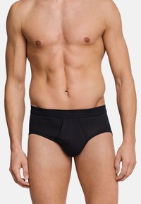 Torso maschile vestito con slip neri a coste, in piedi con le braccia rilassate lungo i fianchi su uno sfondo bianco semplice.