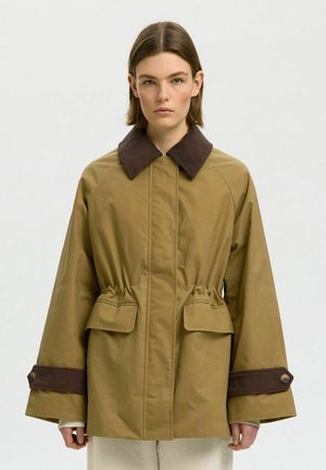 Kobieta w za dużej beżowej kurtce z ciemnobrązowym kołnierzem i mankietami z sztruksu, przednimi kieszeniami z klapkami i taliowanym pasem, stojąca na białym tle.