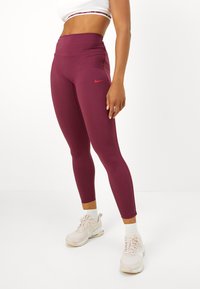 Asmuo vilkintis burgundiškus Nike aukšto liemens legginsus, baltas kojines ir šviesiai rožinius Nike sportbačius, stovintis ant baltų grindų prieš baltą fono.