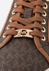 Baskets à motifs bruns avec lacets bruns, œillets dorés et un logo MK doré sur le support de lacet. Tissu texturé avec un accent en cuir lisse.