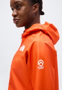 Άτομο με φωτεινό πορτοκαλί μπουφάν North Face Summit Series με κουκούλα, εμφανιζόμενο από τη πλευρά και κομμένο στο ύψος του ώμου και του προσώπου.