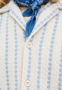 Veste tricotée en crème avec des rayures en zigzag bleu, dotée d'un col pointu et de deux boutons. Associée à un foulard de cou en bandana bleu.